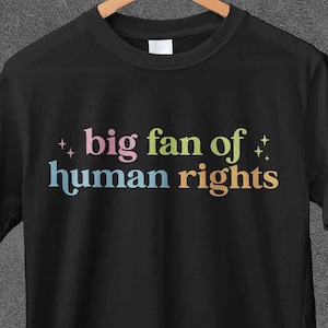 Può includere: T-shirt nero con la scritta multicolore "big fan of human rights". La maglietta è appesa a una gruccia di legno su sfondo grigio. Il marchio "JoJoTeez" è visibile in alto a sinistra.