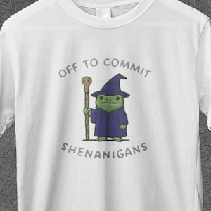 以下が含まれることがあります： 漫画の魔法使いのグラフィックがプリントされた白いTシャツ。緑色の魔法使いは青いローブと帽子を身に着け、杖を持っています。Tシャツのテキストは「Off to Commit Shenanigans」と書かれています。