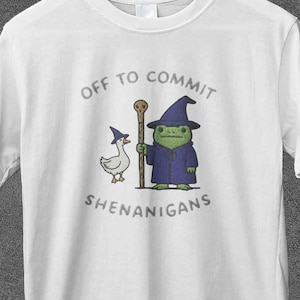 Peut inclure: T-shirt blanc avec un motif fantaisiste. Il présente une grenouille verte magicienne, une oie et le texte "OFF TO COMMIT SHENANIGANS". La grenouille porte une robe et un chapeau violets, tenant un bâton. L'oie porte un chapeau de magicien.
