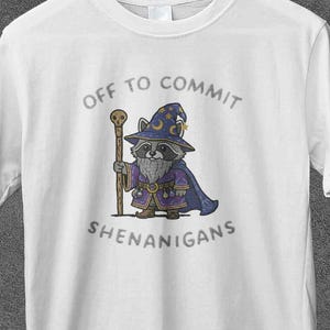 Può includere: T-shirt bianca con un procione dei cartoni animati vestito da mago, con un bastone. Il testo "OFF TO COMMIT SHENANIGANS" è stampato sopra e sotto l'illustrazione. La maglietta è appesa a una gruccia di legno.