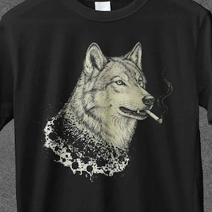 May include: Black t-shirt featuring a detailed illustration of a wolf smoking a cigarette. The wolf is depicted in shades of gray and white, with a dark background. The shirt has the text "JoJoTeez" at the top.