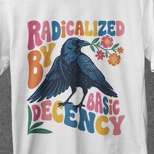 以下が含まれることがあります： 白いTシャツ。暗い青色のカラスが花をくわえているグラフィック。テキスト「RADICALIZED BY BASIC DECENCY」がカラスの上と下にカラフルな文字で印刷されています。木製のハンガーにTシャツがかけられています。