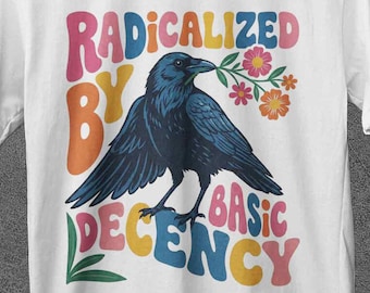 May include: White t-shirt with a graphic of a dark blue crow holding flowers in its beak. The text "RADICALIZED BY BASIC DECENCY" is printed above and below the crow in colorful, retro-style lettering. The shirt is displayed on a wooden hanger.