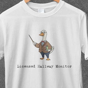 Può includere: T-shirt bianca con un'oca dei cartoni animati con occhiali, papillon e blazer, che tiene un puntatore. Il testo "Licensed Hallway Monitor" è sotto. La maglietta è appesa a una gruccia di legno.