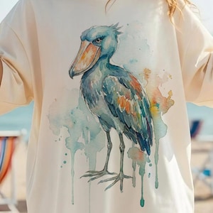 Könnte beinhalten: Cremefarbenes T-Shirt mit einer Aquarellillustration eines Schuhschnabels. Der Vogel ist in Blau-, Grau- und Orangetönen dargestellt, mit einem großen Schnabel und langen Beinen. Das Shirt wird an einem sonnigen Strandtag getragen.