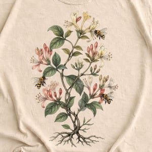 Puede incluir: Camiseta color arena con una ilustración botánica de una planta floreciente con abejas. El diseño incluye elementos rosas, blancos y verdes. La marca "JoJoTeez" está en la parte superior izquierda. También se ve el texto "BULK SALE" y "BUY 3 GET FREE SHIPPING".