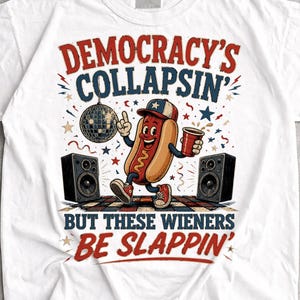May include: A white t-shirt featuring a graphic of a cartoon hot dog wearing a baseball cap, holding a red cup, and dancing between two black speakers. The text reads DEMOCRACY'S COLLAPSIN' BUT THESE WIENERS BE SLAPPIN' with stars and confetti.