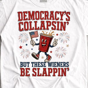 May include: A white short-sleeve t-shirt featuring a graphic of a red plastic cup with a face, arms, and legs, holding an American flag. The text reads DEMOCRACY'S COLLAPSIN' BUT THESE WIENERS BE SLAPPIN' surrounded by red and blue firework illustrations.