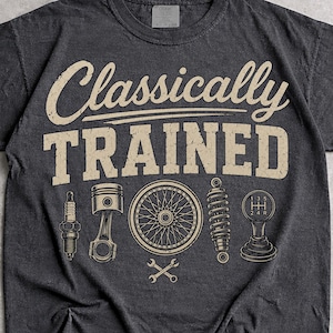 Może przedstawiać: Ciemnoszary t-shirt z krótkim rękawem z napisem Classically TRAINED w kolorze beżowym. Poniżej tekstu znajdują się graficzne ilustracje świecy zapłonowej, tłoka, koła szprychowego, amortyzatora, dźwigni zmiany biegów oraz dwóch skrzyżowanych kluczy płaskich.