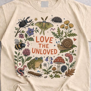 Può includere: Una t-shirt color sabbia con un'illustrazione grafica centrale di vari insetti, una rana, una lumaca e funghi, con il testo "LOVE THE UNLOVED" stampato in lettere arancioni al centro del design circondato da elementi botanici.