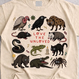 Può includere: Una t-shirt color sabbia con una stampa grafica di vari animali tra cui un avvoltoio, un corvo, una iena, un ratto, un opossum, un serpente, una lucertola, un gatto, un lupo, un alligatore e un ragno, con il testo rosso "LOVE THE UNLOVED".