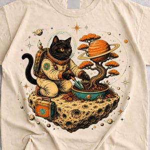 May include: A sand-colored t-shirt featuring a graphic of a black cat in an astronaut suit kneeling on a floating asteroid. The cat is tending to a bonsai tree with a planet-like trunk. The design includes stars, small planets, and a tool box.