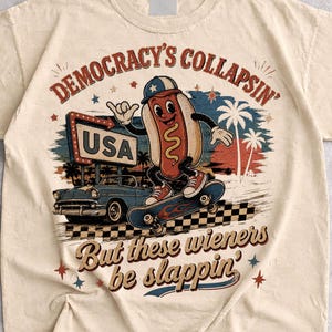 May include: A sand-coloured t-shirt featuring a graphic of a cartoon hot dog on a skateboard. The design includes a vintage car, a sign reading USA, palm trees, and the text Democracy's Collapsin' But these wieners be slappin'.