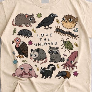 Puede incluir: Una camiseta color arena con un estampado gráfico de varios animales, incluyendo una rata, un cuervo, un pez globo, un buitre, una zarigüeya, un mapache, un pez gota, una mofeta y un calamar. El texto "LOVE THE UNLOVED" aparece impreso en el centro.