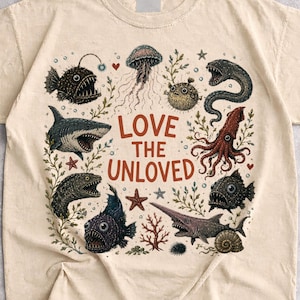 Peut inclure: Un t-shirt à manches courtes de couleur sable avec un motif central représentant diverses créatures des abysses, dont un requin, une méduse et un calmar. Le texte 'LOVE THE UNLOVED' est imprimé au centre en rouge-brun.