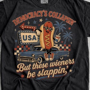 May include: A black t-shirt featuring a graphic of a hot dog wearing sunglasses, leaning against a blue vintage car. The text reads "DEMOCRACY'S COLLAPSIN" and "But these wieners be slappin'" with a "USA" sign and palm trees in the background.