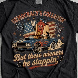 May include: A black short-sleeve t-shirt featuring a graphic of a cartoon hot dog driving a blue muscle car. The design includes a stylised American flag background, fireworks, and the text "DEMOCRACY'S COLLAPSIN' But these wieners be slappin'" in a vintage-style font.