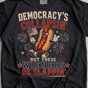 May include: A black short-sleeve t-shirt featuring a graphic of a hot dog character wearing sunglasses and holding an American flag. The text reads DEMOCRACY'S COLLAPSIN' BUT THESE WIENERS BE SLAPPIN' surrounded by red, white, and blue firework designs.