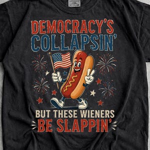 May include: A black short-sleeve t-shirt featuring a graphic of a cartoon hot dog holding an American flag. The text reads DEMOCRACY'S COLLAPSIN' BUT THESE WIENERS BE SLAPPIN' with red and blue lettering and firework illustrations in the background.