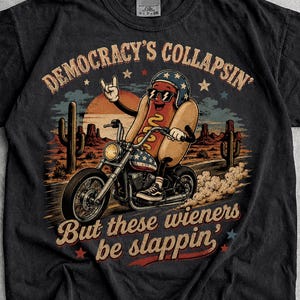 May include: A black t-shirt featuring a graphic of a hot dog wearing sunglasses and a helmet, riding a motorcycle through a desert landscape with cacti. The text reads, "DEMOCRACY'S COLLAPSIN' But these wieners be slappin'" in a vintage-style font.