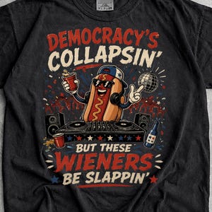 May include: A black short-sleeve t-shirt featuring a graphic of a hot dog wearing sunglasses and a baseball cap, DJing with a mixer and speakers. The text reads: DEMOCRACY'S COLLAPSIN' BUT THESE WIENERS BE SLAPPIN'. The design includes red and blue stars.