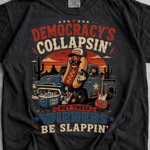 May include: A black short-sleeve t-shirt featuring a graphic of a hot dog character wearing sunglasses and a cap, leaning against a blue pickup truck. The text reads "DEMOCRACY'S COLLAPSIN' BUT WIENERS BE SLAPPIN'" with a guitar and cooler visible.