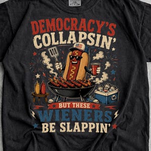 May include: A black short-sleeved t-shirt featuring a graphic of a hot dog wearing sunglasses and a baseball cap, grilling sausages. The text reads: DEMOCRACY'S COLLAPSIN' BUT THESE WIENERS BE SLAPPIN'. The design includes red, white, and blue accents.
