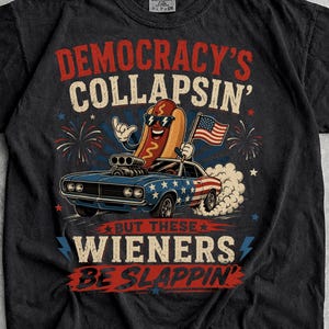 May include: A black t-shirt featuring a graphic of a cartoon hot dog wearing sunglasses, driving a blue muscle car with an American flag pattern. The text reads DEMOCRACY'S COLLAPSIN' BUT THESE WIENERS BE SLAPPIN' in red and white lettering.