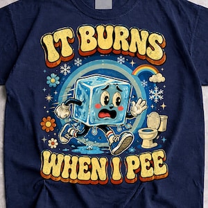 Może przedstawiać: Granatowy t-shirt z grafiką przedstawiającą kreskówkową kostkę lodu z twarzą biegnącą w stronę toalety. Napis "IT BURNS" znajduje się nad kostką lodu, a "WHEN I PEE" poniżej, wykonany żółtą, postarzaną czcionką w stylu retro.