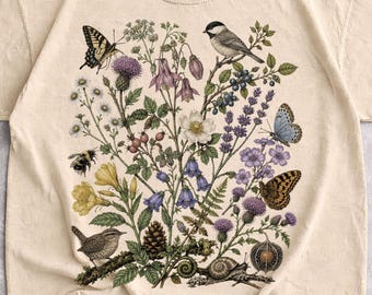 May include: A sand-colored short-sleeve t-shirt featuring a detailed botanical illustration. The graphic displays various wildflowers, a small bird, a bumblebee, a snail, and several butterflies in muted natural tones across the center chest.