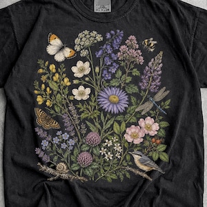 Könnte beinhalten: Ein schwarzes Kurzarm-T-Shirt mit einer detaillierten botanischen Illustration verschiedener Wildblumen, einem Schmetterling, einer Libelle, einer Schnecke und einem kleinen Vogel. Das Motiv ist mittig auf der Brust platziert und zeigt violette, gelbe und rosa Blüten.