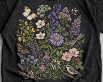 May include: A black short-sleeve t-shirt featuring a detailed botanical illustration of various wildflowers, a butterfly, a dragonfly, a snail, and a small bird. The graphic is centered on the chest and includes purple, yellow, and pink floral elements.