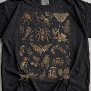 Könnte beinhalten: Ein schwarzes Kurzarm-T-Shirt mit einem detaillierten, hellbraunen Grafikdruck verschiedener Insekten, Spinnen und botanischer Elemente. Das Design zeigt Käfer, einen Falter, eine Spinne, eine Gottesanbeterin und Pilze in einer zentralen Anordnung.