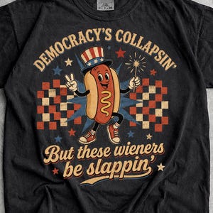 May include: A black t-shirt featuring a graphic of a cartoon hot dog wearing a patriotic top hat, holding a sparkler, and making a peace sign. The text reads "DEMOCRACY'S COLLAPSIN' But these wieners be slappin'" surrounded by red and blue stars.