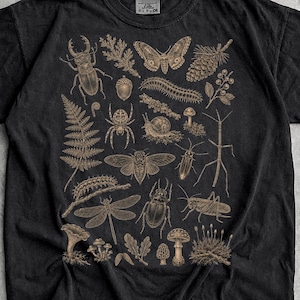 Op de afbeelding: Een zwart t-shirt met korte mouwen en een gedetailleerde beige grafische print van diverse insecten, spinnen, slakken, paddenstoelen, varens en dennenappels, gerangschikt in een centraal cluster op de voorzijde van het kledingstuk.