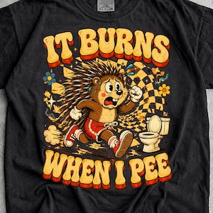Può includere: Una t-shirt nera con la grafica di un riccio dei cartoni animati che corre verso un water. Il testo IT BURNS WHEN I PEE è scritto in grandi lettere arancioni e gialle. Il design include un motivo a scacchi sullo sfondo e piccoli fiori.