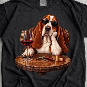 Op de afbeelding: Een zwart t-shirt met korte mouwen en een afbeelding van een bruin-witte Basset Hound met een zwarte zonnebril. De hond rust met zijn kin op een poot aan een ronde houten tafel met een glas rode wijn, een kurkentrekker en twee wijnkurken.