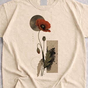 Pode incluir: Uma t-shirt cor de areia com uma impressão gráfica de uma papoila vermelha, uma forma circular escura, uma linha vertical e um bloco retangular com salpicos de tinta preta, centrada na parte frontal da peça de vestuário de manga curta.