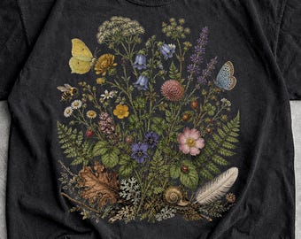 May include: A black short-sleeve t-shirt featuring a detailed botanical illustration on the front. The graphic displays a colorful arrangement of various wildflowers, green ferns, a yellow butterfly, a blue butterfly, a bee, and a snail.