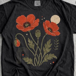 May include: A black short-sleeve t-shirt featuring a graphic print of two large red poppy flowers, a poppy bud, green leaves, a beige circle, and a thin line arch. The design is centered on the chest of the garment, which has a crew neckline.