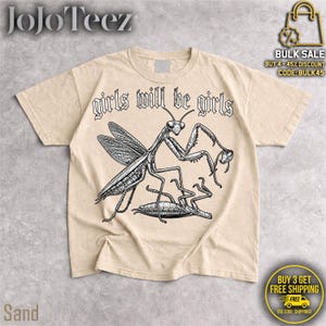 May include: A sand-colored t-shirt featuring a black and white line art illustration of a praying mantis standing over another insect. The text "girls will be girls" is printed in a gothic-style font above the insect graphic on the front.