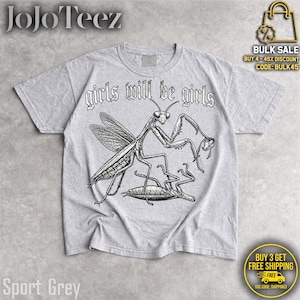 May include: A light gray short-sleeve t-shirt featuring a black ink-style illustration of a praying mantis standing over another insect. The text "girls will be girls" is printed in a gothic-style font above the graphic on the front of the shirt.
