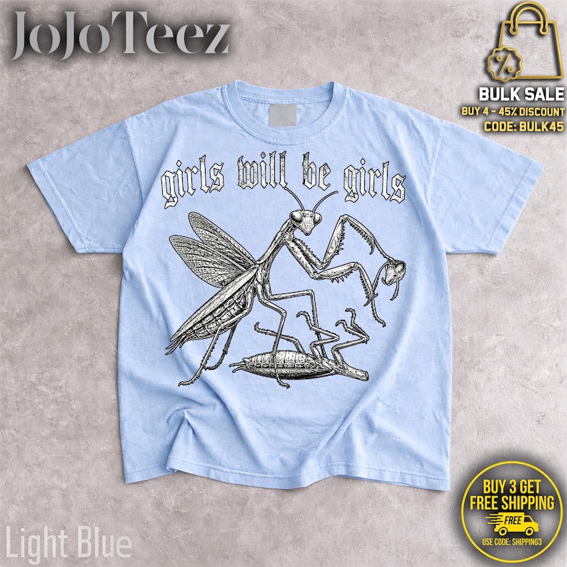 May include: A light blue t-shirt featuring a black and white graphic illustration of a praying mantis standing over another insect. The text "girls will be girls" is printed in a stylized black font above the insect illustration.