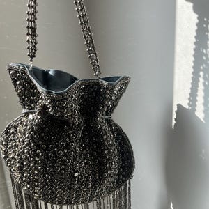 May include: A dark grey drawstring pouch bag covered in intricate metallic beadwork and sequins. The bag features a long beaded fringe along the bottom edge and a matching beaded shoulder strap. The interior is lined with a smooth grey fabric.