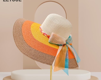 Pode incluir: Um chapéu de palha de abas largas com um design colorido. O chapéu apresenta uma copa creme e uma aba com listras marrons, laranja e amarelas. Uma fita colorida e um lenço estampado são amarrados ao redor do chapéu. A palavra "hat" e a marca "ZEYGUL" são impressas no canto superior esquerdo.