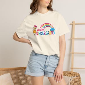 May include: A cream-colored t-shirt featuring a graphic of a smiling pencil, a colorful rainbow, and a blue cloud. The text reads Hello KINDERGARTEN in multi-colored letters. The shirt has short sleeves and a crew neckline, paired with light blue denim shorts.