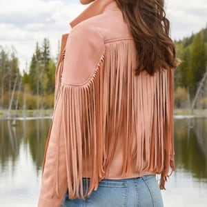 Könnte beinhalten: Hellrosa Wildlederjacke mit langen Fransen am Rücken und an den Ärmeln. Die Jacke hat einen klassischen Kragen und eine taillierte Passform. Das Model trägt eine blaue Jeans. Der Hintergrund zeigt einen See und Bäume.