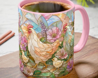 May include: A pink-handled coffee mug featuring a stained-glass design of chickens, chicks, and flowers. The mug is filled with dark coffee and sits on a wooden cutting board. The background includes pine cones and greenery.