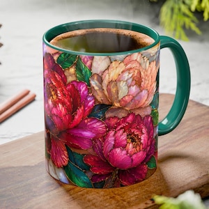 May include: A ceramic mug with a vibrant floral design featuring red, pink, and peach peonies. The mug has a green handle and rim, and is filled with a dark liquid. The mug is placed on a wooden cutting board.
