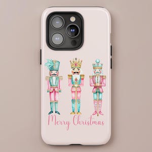 May include: A phone case with a light pink background featuring three watercolor nutcracker soldiers in pink, teal, and gold. The words "Merry Christmas" are written in pink script. The brand name "LILOU" is at the bottom.
